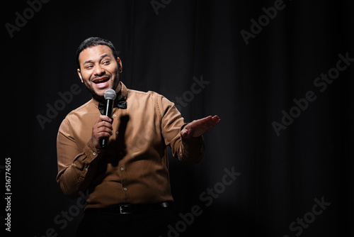 cheerful indian comedian in shirt and bow tie holding microphone during monologue on black.