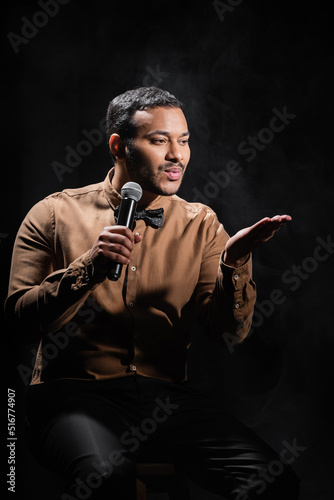 indian comedian sitting and pouting lips while performing stand up comedy into microphone on black.