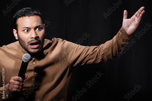 shocked indian stand up comedian telling joke into microphone on dark stage isolated on black.