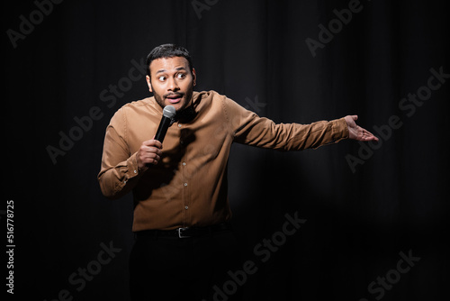 puzzled indian comedian in shirt and bow tie holding microphone and gesturing during monologue on black. photo