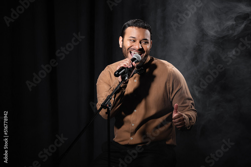 Fototapeta happy indian comedian sitting on chair and performing stand up comedy into microphone on black with smoke