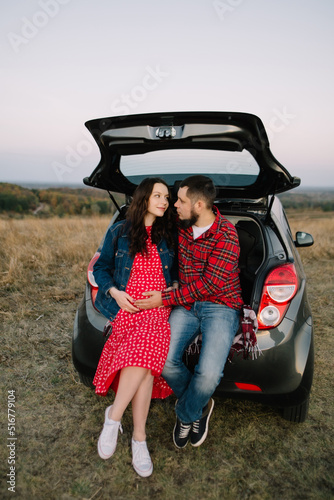 Young happy family sits in the trunk of a new car and cheerfully communicates. Mom-to-ne and dad-to-be hug and gently hold on to their stomach belly while waiting for the baby. Vertical photo