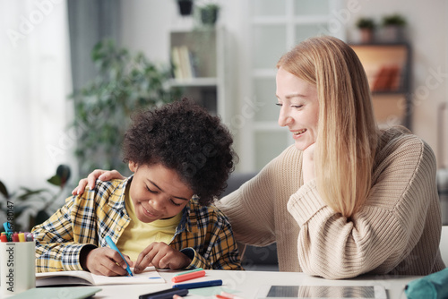 Young mother sitting at table in the room with her adopted son while he drawing with crayons photo