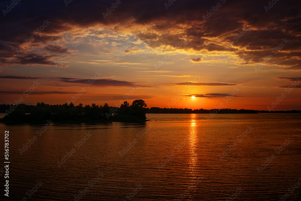 Sunset over the river (lake, bay). Forest in the distance. Orange clouds. The light of the sun is reflected in the water. Waves on the surface of the water