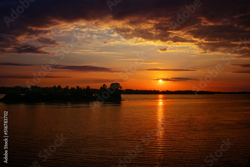 Sunset over the river (lake, bay). Forest in the distance. Orange clouds. The light of the sun is reflected in the water. Waves on the surface of the water © Vlad Kazhan