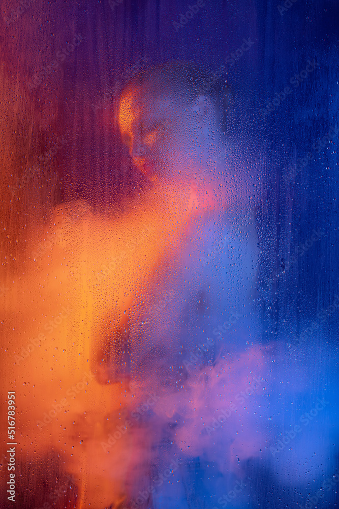 Portrait of young woman with bright makeup posing isolated over blue background in neon light. Foggy glass and smoke effect