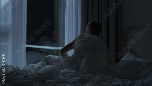 Handsome young adult slowly wakes up early in a cozy apartment, checks his watch and gets out of bed. Young and Fit caucasian Man greeting new day. Early morning before sunrise.