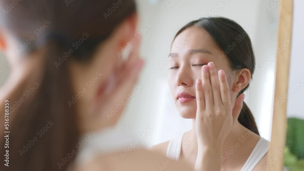 Young Asian woman apply serum and moisturizing cream on healthy facial skin and look at mirror Young Asian woman apply serum and moisturizing cream on healthy facial skin and look at mirror