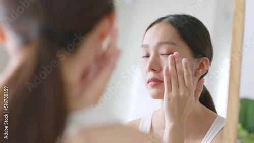 Young Asian woman apply serum and moisturizing cream on healthy facial skin and look at mirror