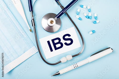 On a blue background, a stethoscope, a medical mask, pills, a pen and a white card with the text IBS Irritable Bowel Syndrome. View from above.