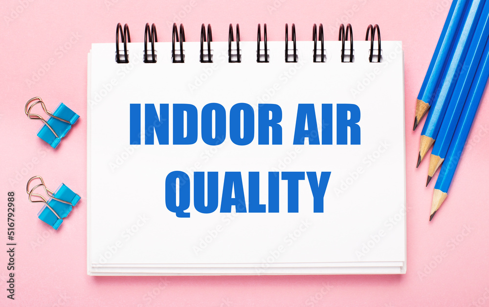 On a light pink background, light blue pencils, paper clips and a white notebook with the text INDOOR AIR QUALITY On a light pink background, light blue pencils, paper clips and a white notebook with the text INDOOR AIR QUALITY
