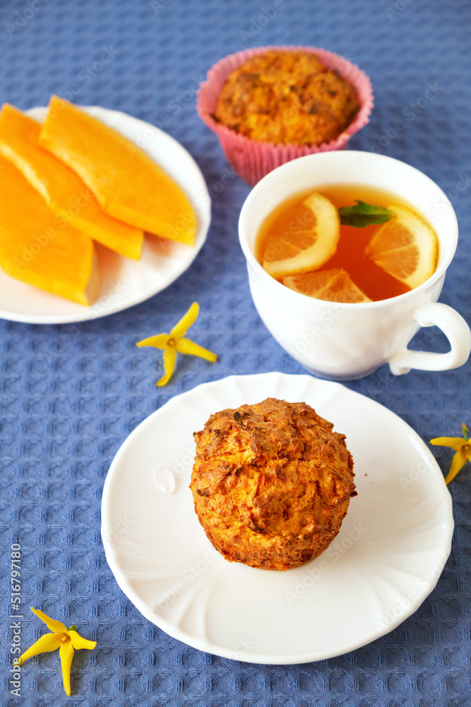 Homemade baked pastries. Healthy pumpkin muffins with traditional fall spices, pieces of pumpkin on saucer. White cup with tea on blue tablecloth. Selective focus. Vertical Homemade baked pastries. Healthy pumpkin muffins with traditional fall spices, pieces of pumpkin on saucer. White cup with tea on blue tablecloth. Selective focus. Vertical