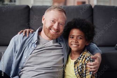 Portrait of dad and adopted son embracing and smiling at camera sitting at home photo
