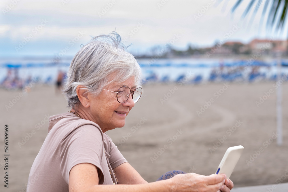Caucasian senior woman sitting at the beach in a summer cloudy day using mobile phone, attractive female enjoying relax and holiday