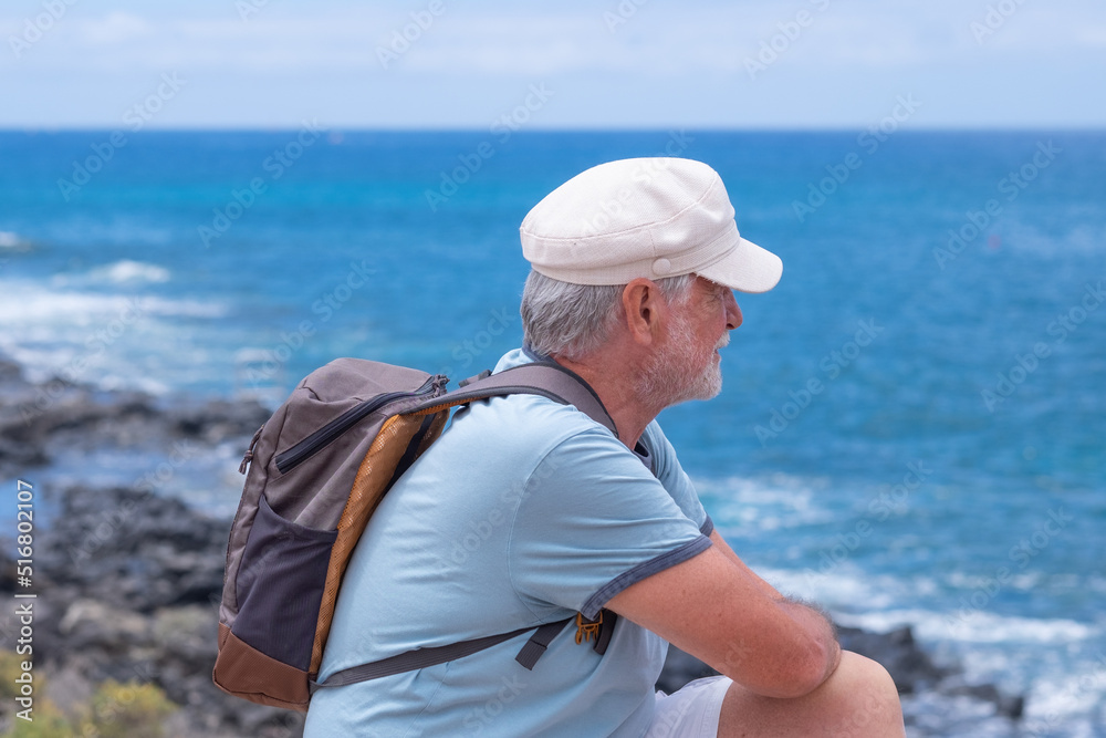 Smiling caucasian bearded senior man with hat sitting at the beach looking at the horizon while enjoying vacation or retirement. Horizon over the water, copy space