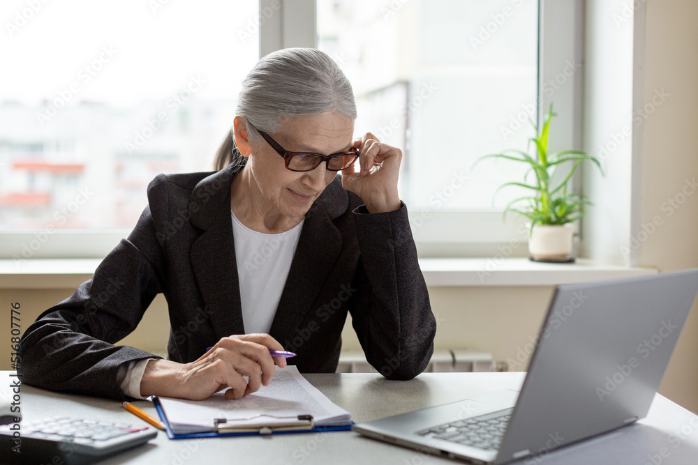 Intelligent caucasian senior woman in formal wear adjust her glasses and look critically at laptops screen with a pen in hand, indoor, selective focus.