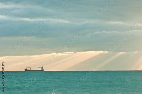 front view, very far distance of a cargo ship motoring tropical waters after a rain storm, with a shard of sunlight, cutting through the clouds
