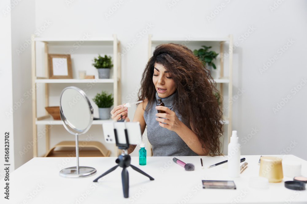 Makeup artist. Lovely tanned handsome curly Latin beauty blogger in casual top present serum for skin rejuvenation in home interior. Copy space. Influencer record video review blog using smartphone Makeup artist. Lovely tanned handsome curly Latin beauty blogger in casual top present serum for skin rejuvenation in home interior. Copy space. Influencer record video review blog using smartphone