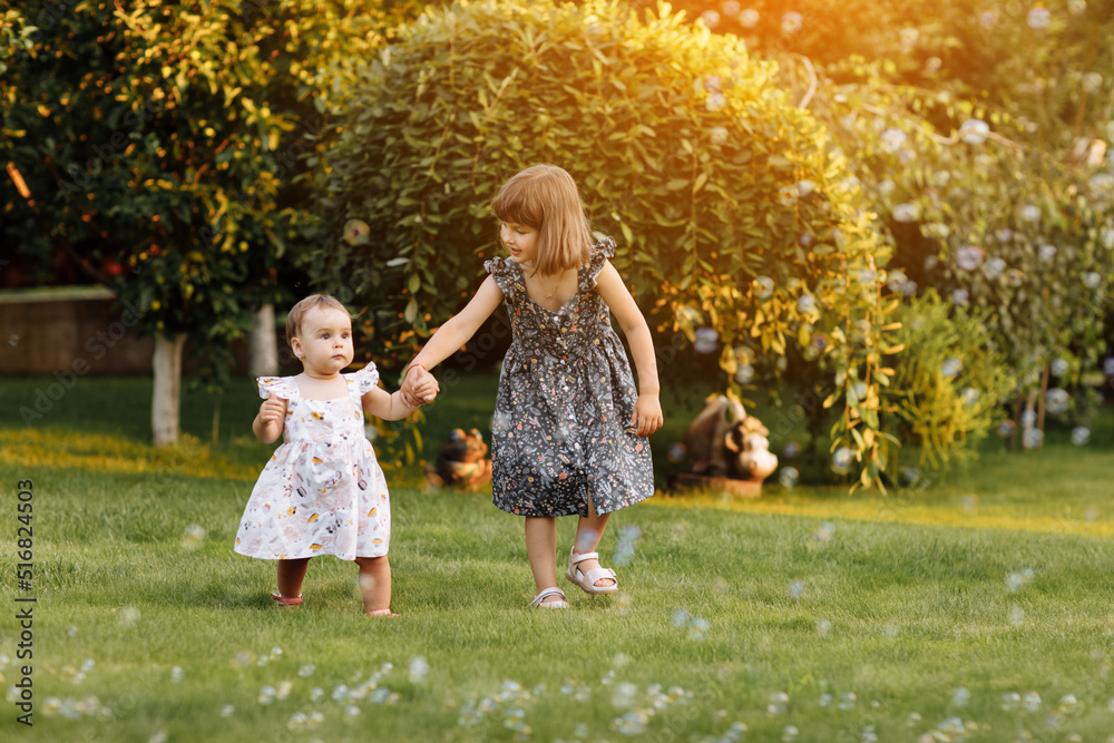 Two happy girls, adorable sisters are smiling happily together. Child girl holding hand her younger baby sister and playing outdoors on summer day holiday vacation, best friends.Family day Two happy girls, adorable sisters are smiling happily together. Child girl holding hand her younger baby sister and playing outdoors on summer day holiday vacation, best friends.Family day