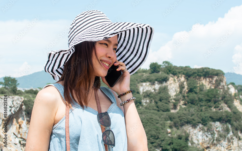 Beautiful Asian female traveler wearing casual fashionable clothes, hat, talking on mobile phone, standing on the top of mountains, smiling with happiness while traveling in summer holiday vacation.