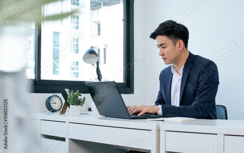 Asian handsome business man wearing white shirt and blue formal suit, communicating, working, typing on laptop, sitting near window in indoor office and smiling with happiness.