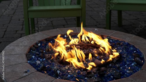 Yellow flames dance over a gas or propane powered circular fire pit with blue rocks and relaxing chairs. Hand held with zoom in.