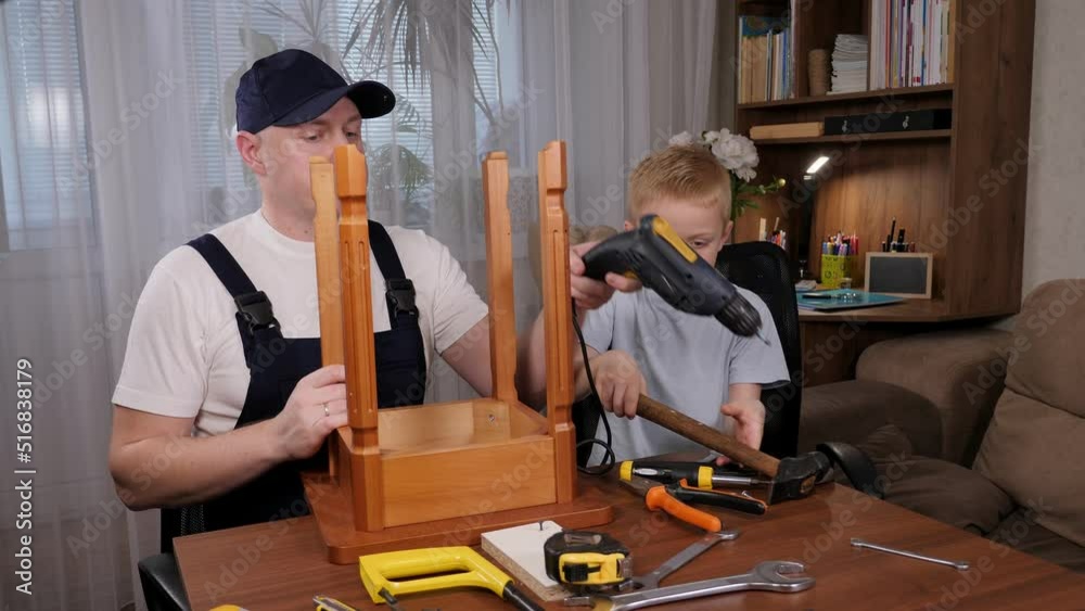 An adult builder father in overalls teaches his son how to repair a wooden chair, and shows the boy how to use an electric drill. There are many different tools on the table. An adult builder father in overalls teaches his son how to repair a wooden chair, and shows the boy how to use an electric drill. There are many different tools on the table.