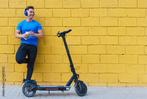 Young Caucasian boy smiling with headphones, blue t-shirt and with an electric scooter leaning against a yellow wall.