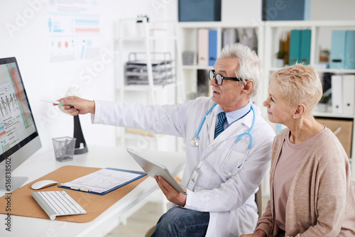 Confident senior male practitioner with gray hair sitting at table and pointing at computer screen while consulting mature female patient in clinic