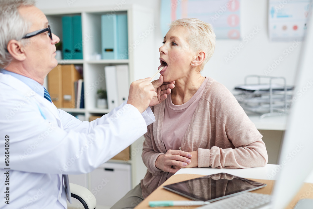 Fototapeta premium Serious male otolaryngologist in white coat sitting at table and examining throat of sick senior patient in own office