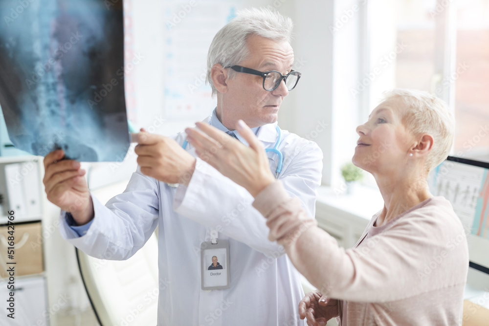Obraz premium Attractive blond-haired mature lady pointing at core x-ray image while discussing her pain with doctor