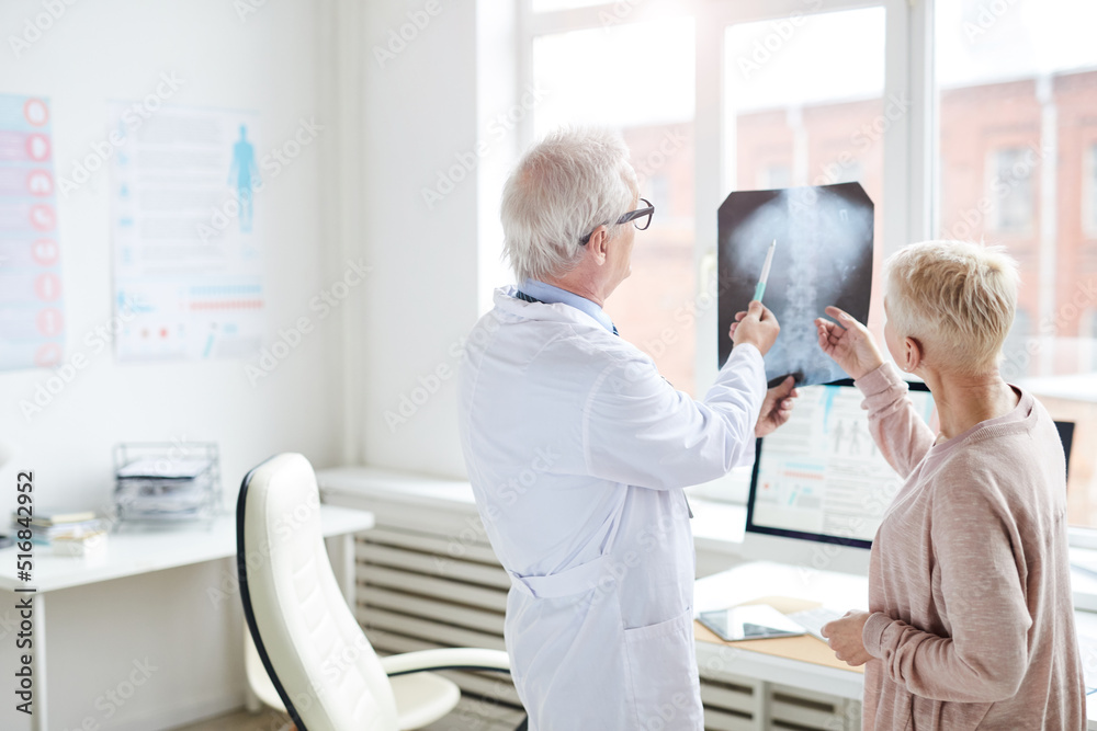 Obraz premium Rear view of gray-haired doctor in lab coat standing against window and watching x-ray image of spinal cord with female patient
