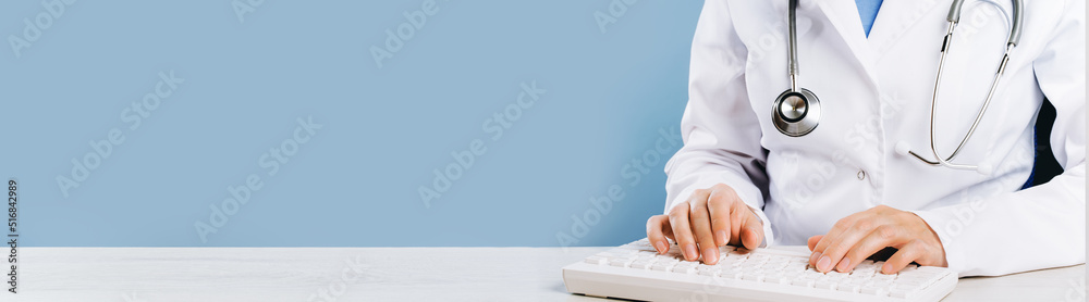Doctor typing on computer keyboard in office. Physician in uniform uses computer at desk. Professional diagnosis and treatment tele medicine. Professional healthcare wide banner - obrazy, fototapety, plakaty 