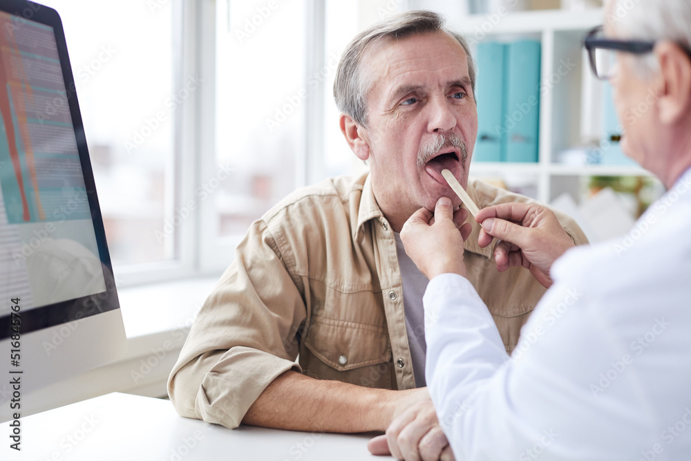 Over shoulder view of male doctor sitting at table and using tongue depressor while looking at throat of senior patient Over shoulder view of male doctor sitting at table and using tongue depressor while looking at throat of senior patient