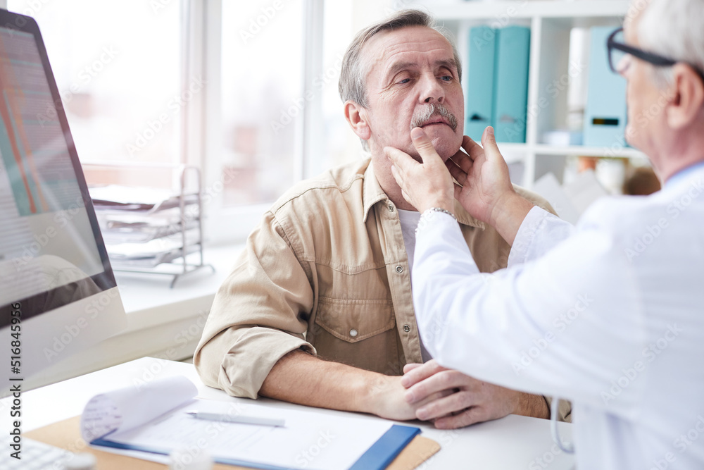 Over shoulder view of senior doctor sitting at table and touching neck of mature man while doing neck lump examination Over shoulder view of senior doctor sitting at table and touching neck of mature man while doing neck lump examination