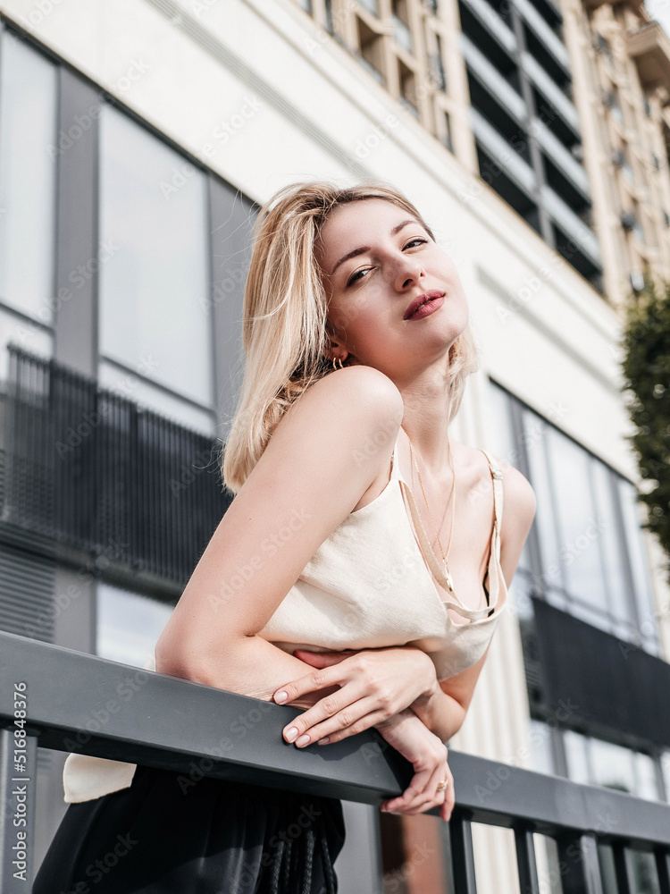 Street shot of a blonde girl in a pink shirt and black pants, against the background of a new building Street shot of a blonde girl in a pink shirt and black pants, against the background of a new building