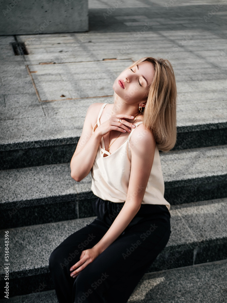Street shot of a blonde girl in a pink shirt and black pants, against the background of a new building