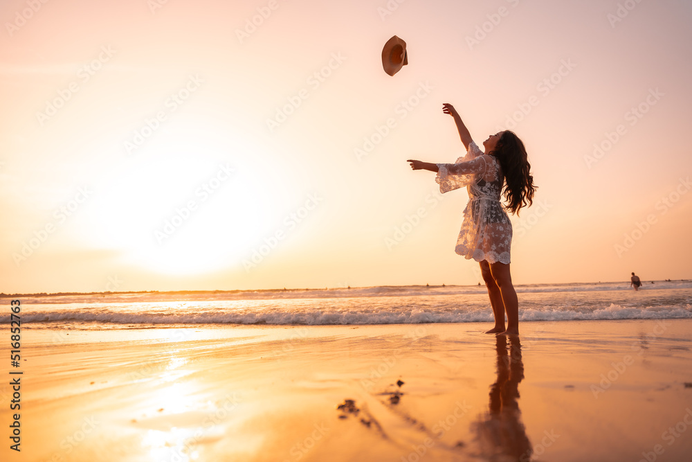 A woman in the sunset in a white dress at the sea in summer throwing the hat on vacation