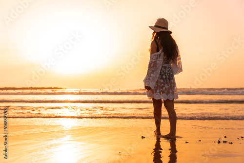 A woman at sunset in a white dress with a hat walking by the sea at low tide