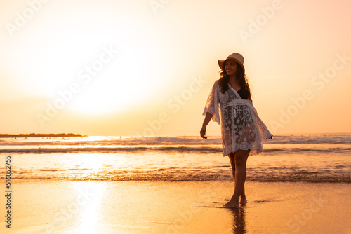 A woman in the sunset in a white dress with a hat walking by the sea