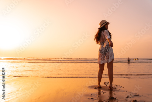 A woman in the sunset in a white dress at the sea in summer with a hat