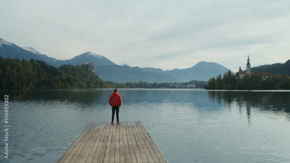 Young man standing alone on lake footbridge and admires sunrise on a lake Bled, Slovenia. Early chilly morning.