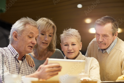 Group of positive retired friends in casual clothing sitting at table and using digital tablet while watching video online together