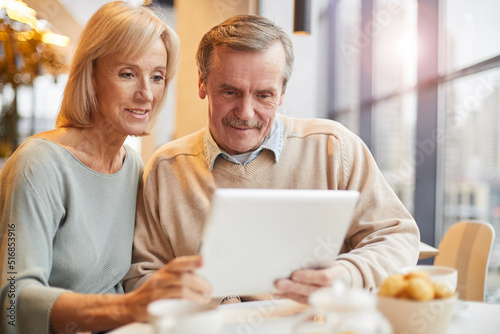 Smiling senior couple in casual clothing sitting at table and using modern device while surfing net together in cafe