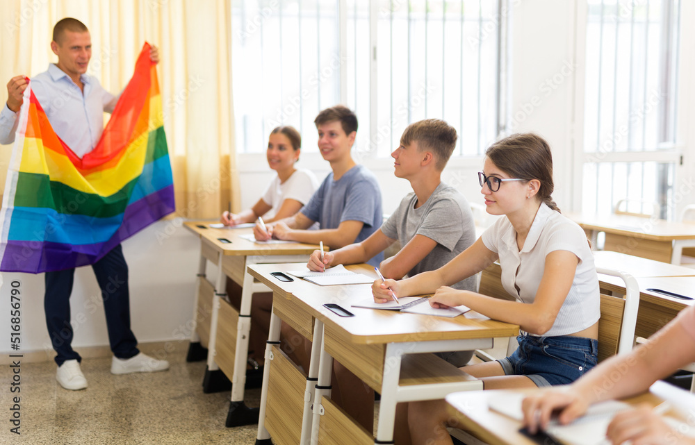 Focused teenage schoolgirl sitting at lesson with classmates, listening young teacher talking about LGBT community and showing rainbow flag