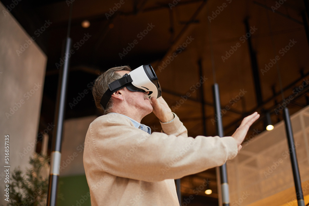 Below view of concentrated senior man in virtual reality goggles gesturing hand while playing video games Below view of concentrated senior man in virtual reality goggles gesturing hand while playing video games