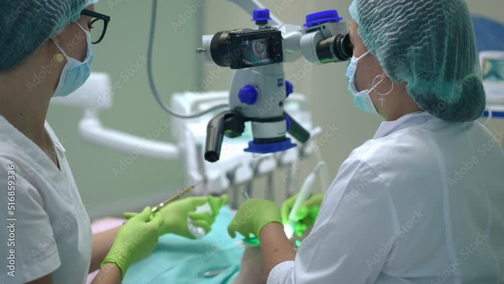 Concentrated dentist examining tooth hole with dental microscope as assistant passing tools in slow motion. Caucasian professional orthodontist and nurse curing patient in dental chair in hospital