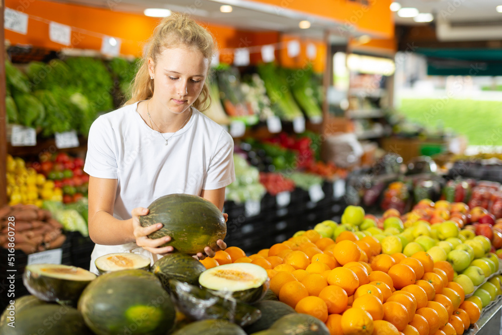 Focused fifteen-year-old girl, who came to the supermarket for shopping, chooses a ripe juicy melon, standing at the counter