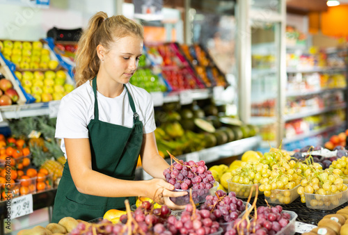 Fotografie Fifteen-year-old girl who works part-time in a store as a trainee saleswoman puts ripe grapes on the counter, laid out