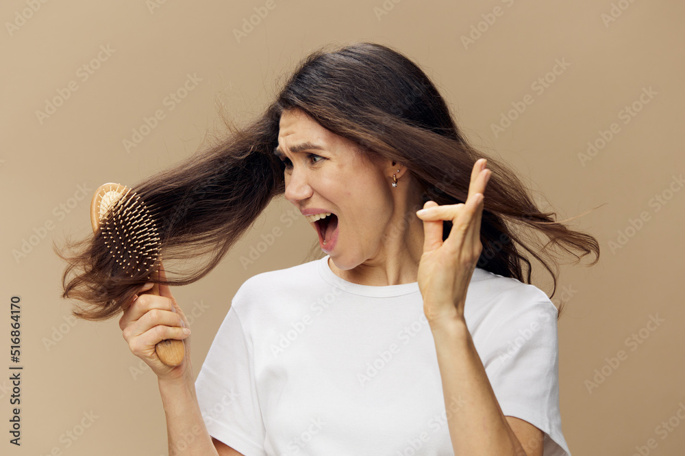 Fototapeta premium A sad, upset woman combs her hair with a wooden massage comb and screams, showing the fallen hair to the camera. Horizontal photo on a beige background.